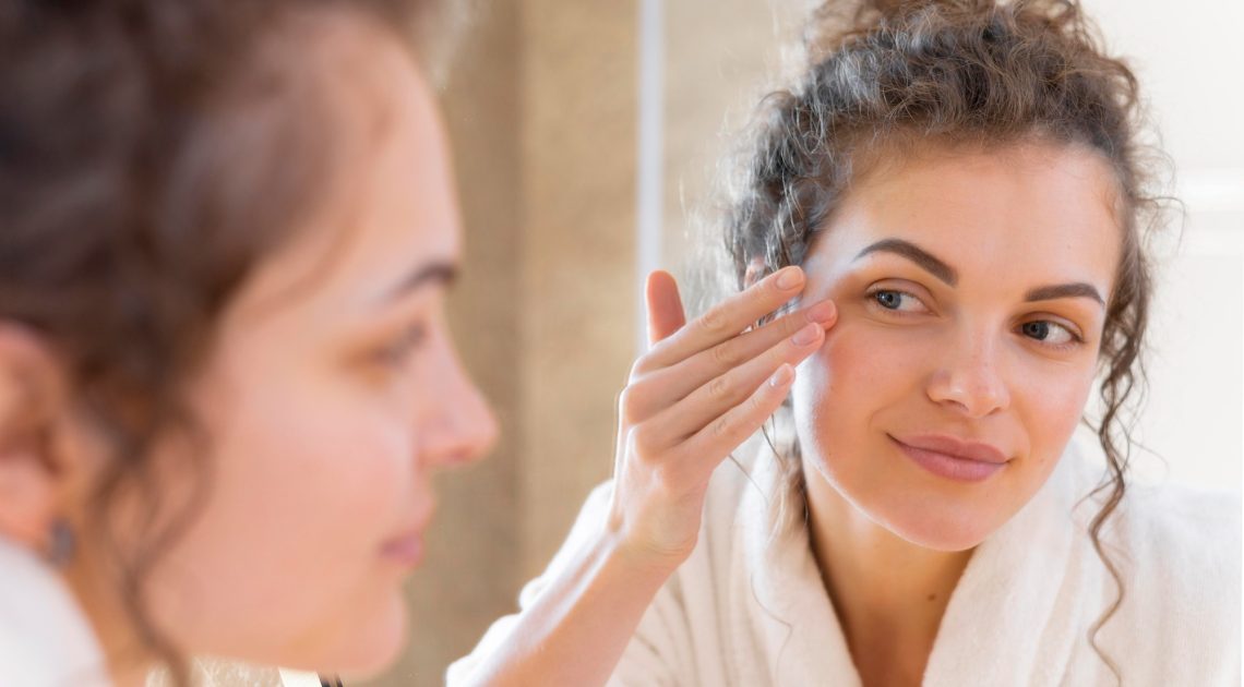 woman-applying-cream-face-while-looking-mirror